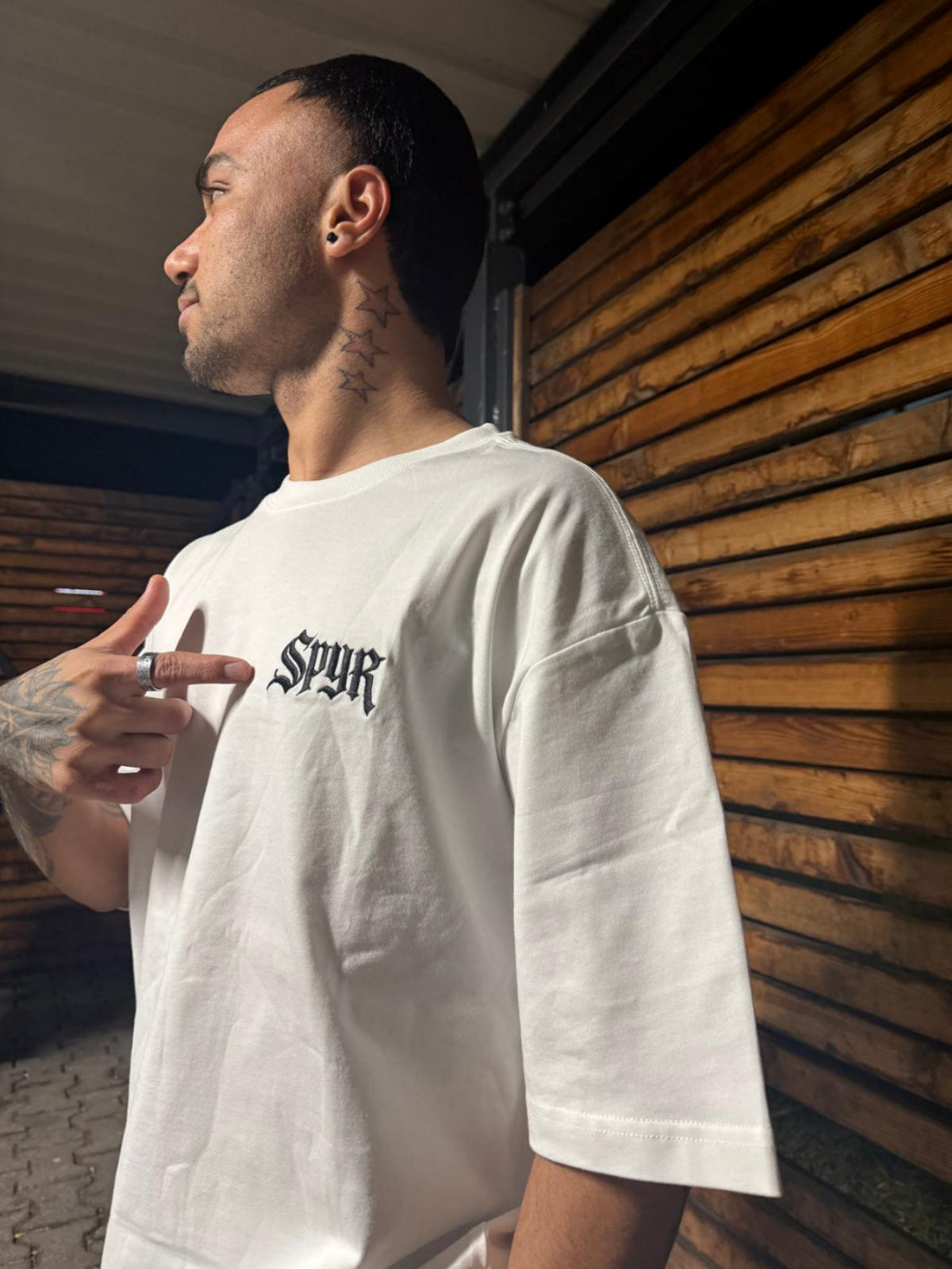 Man wearing a white t-shirt with 'SPYR' logo against a wooden wall.