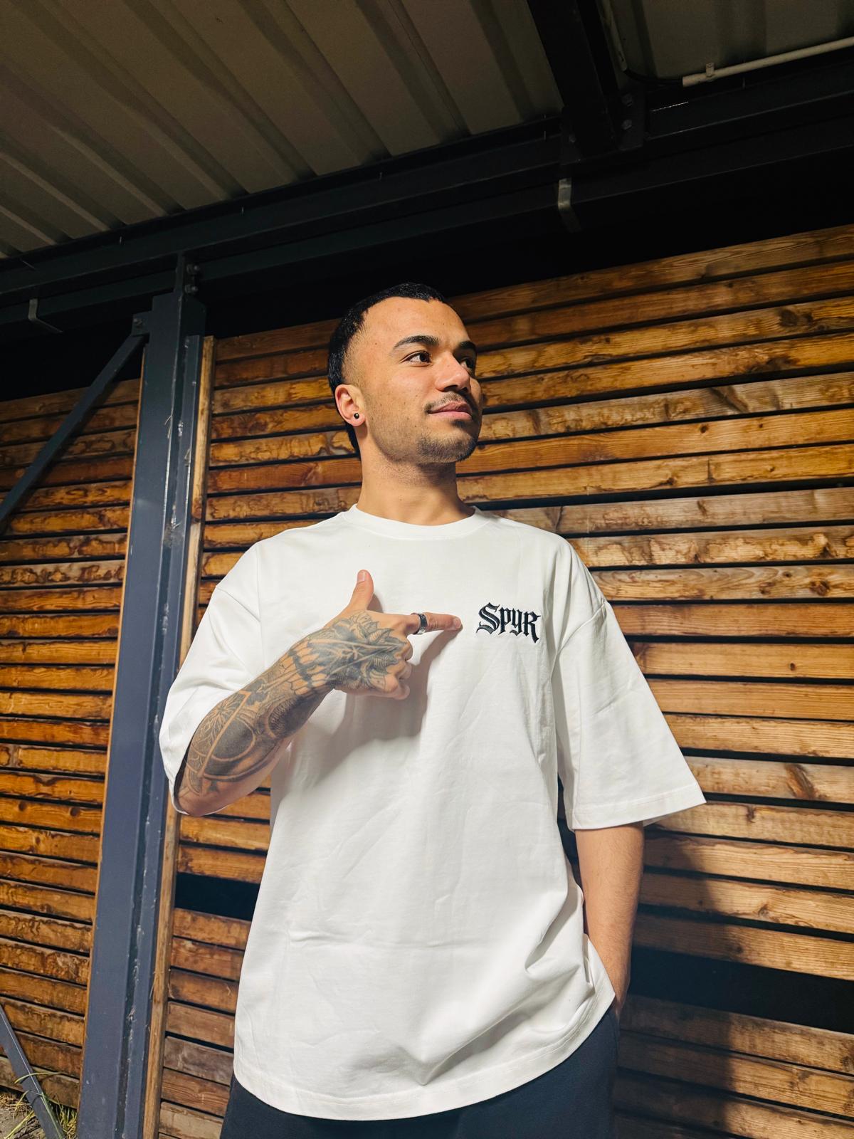 Man wearing a white t-shirt with a logo, standing against a wooden wall.