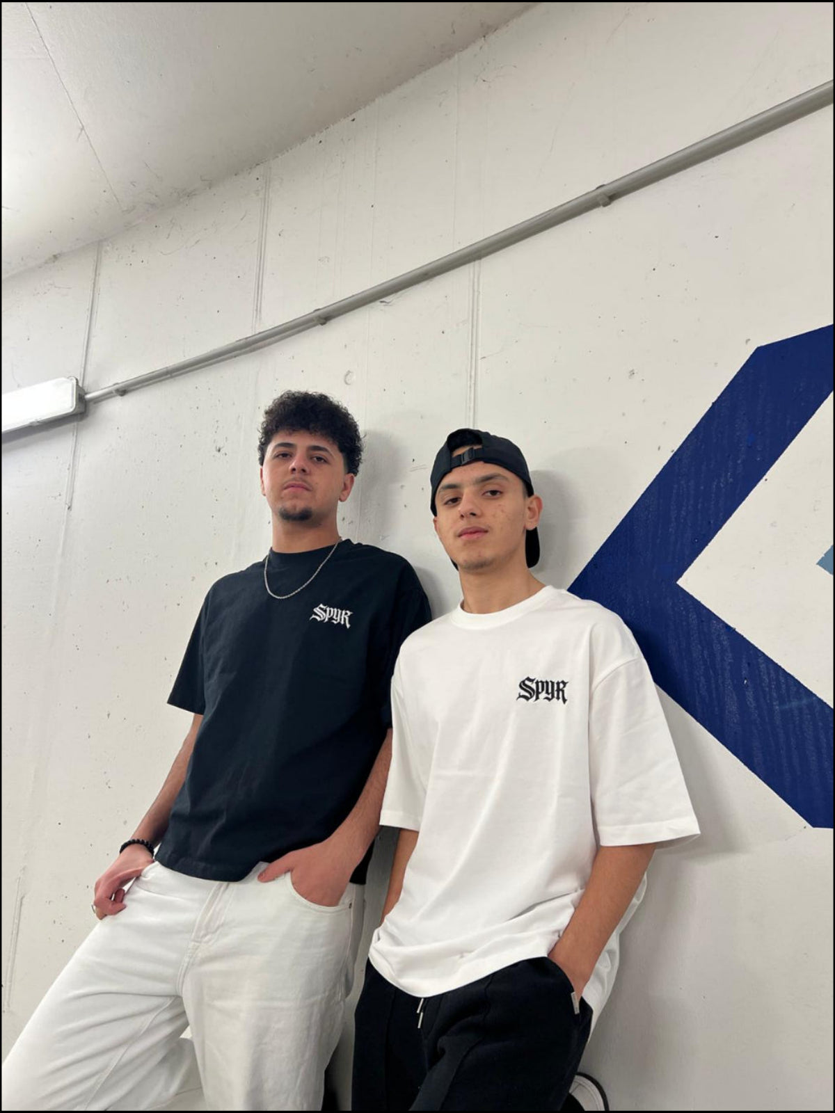 Two people wearing black and white t-shirts with a logo, standing against a white wall with a blue logo.