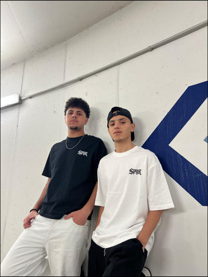 Two people wearing black and white t-shirts with a logo, standing against a white wall with a blue logo.